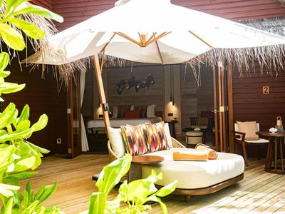 Liegestuhl mit bunten Kissen unter einem Sonnenschirm auf einer Holzterrasse neben grüner Pflanze.