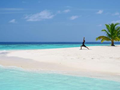 Frau macht Yoga auf einem weißen Sandstrand mit Palmen und türkisfarbenem Wasser.