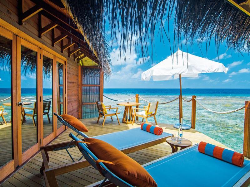 Terrasse mit Liegestühlen und Tischen direkt am Meer unter blauem Himmel.