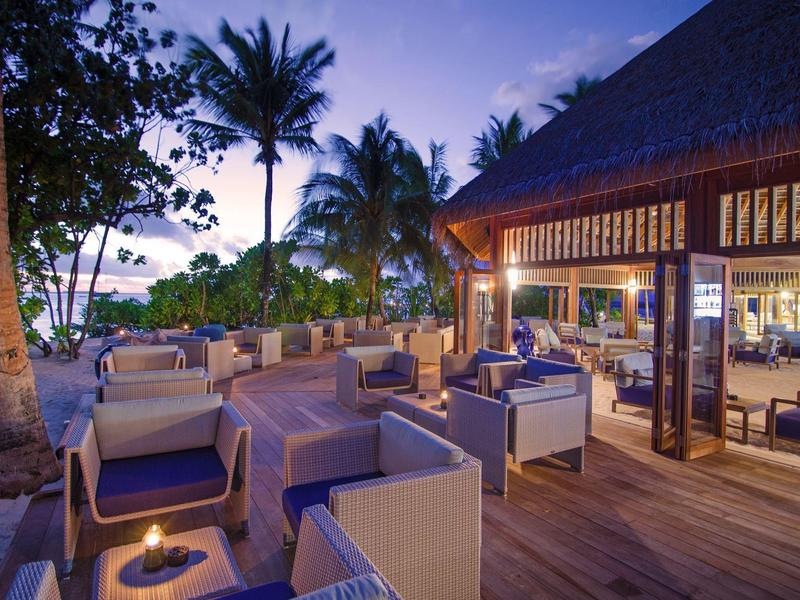 Gemütliche Außenlounge mit Sesseln und Tischen, Palmen am Strand bei Abenddämmerung.