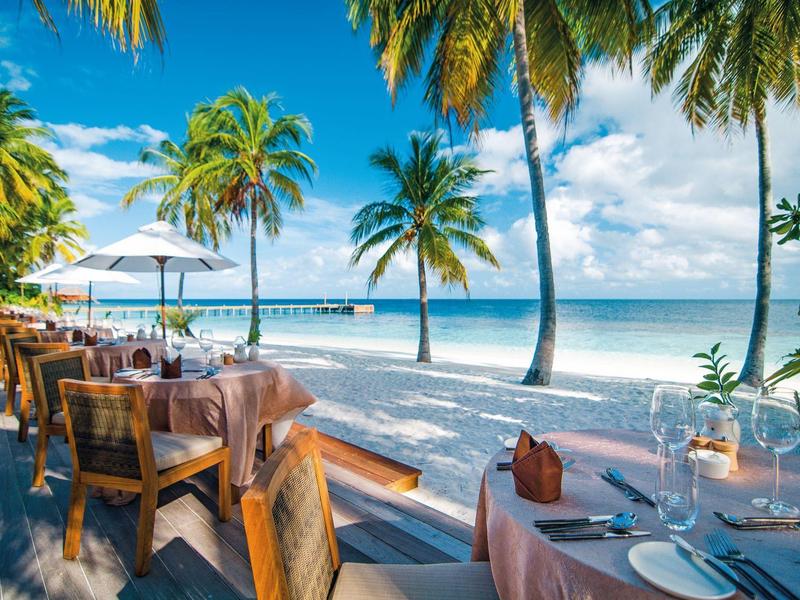 Restauranttische am Strand mit Blick auf das Meer und Palmen bei sonnigem Wetter.