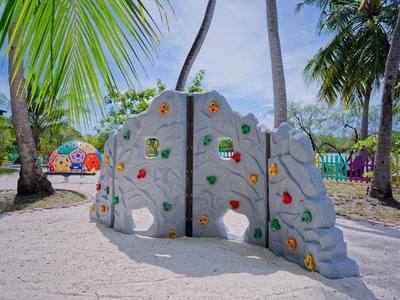 Kleiner Spielplatz mit buntem Kletterfelsen, Palmen und Sandboden unter blauem Himmel.