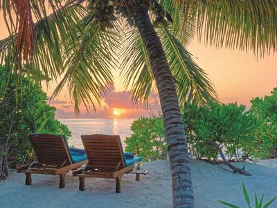 Zwei Liegestühle am Strand unter Palmen bei Sonnenuntergang über dem Meer.