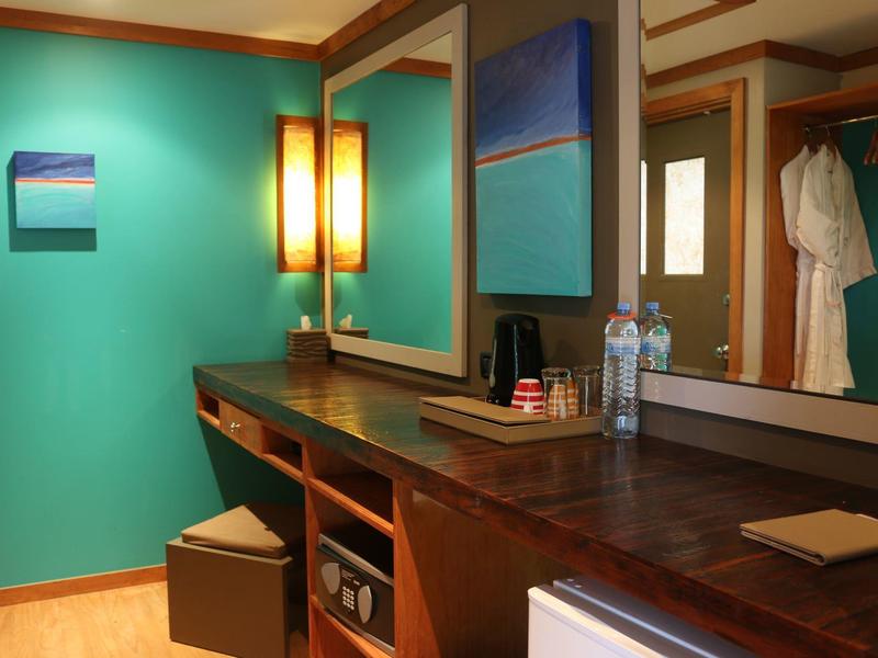 Ein heller Raum mit türkisfarbener Wand, Holztisch, Spiegel, zwei Lampen und kleinen Kunstwerken.