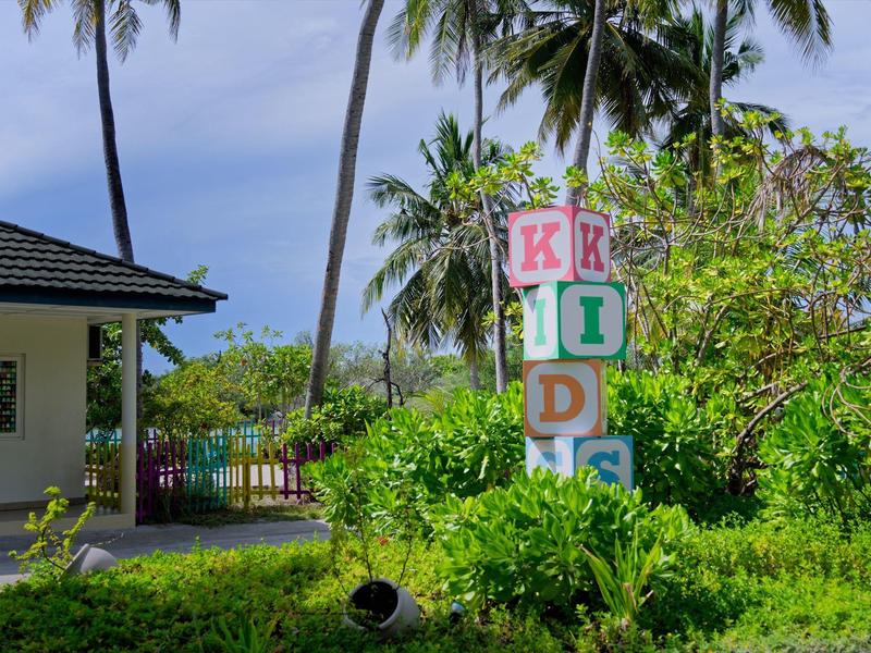 Bunte Kinderspielplatz-Schilder zwischen Palmen und grünen Pflanzen bei bewölktem Himmel.