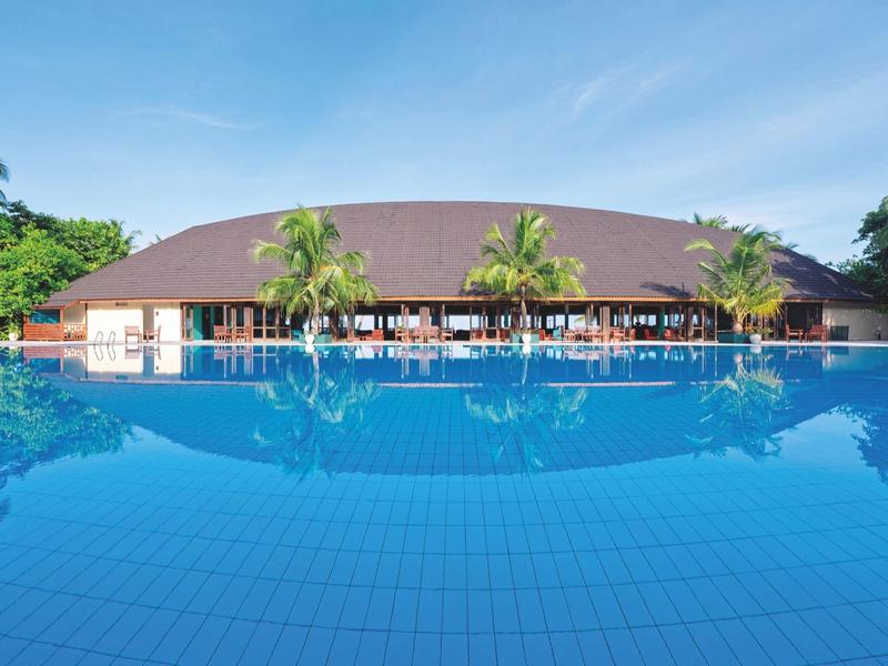 Großer Pool mit klarem Wasser vor rundem Gebäude und Palmen, blauer Himmel im Hintergrund.