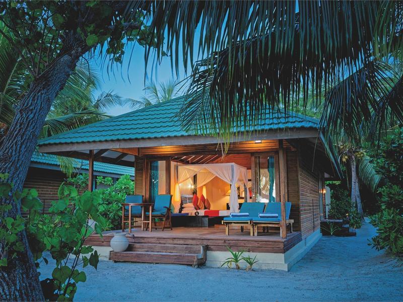 Holzhütte mit Veranda, umgeben von Palmen, beleuchtet bei Dämmerung am Sandstrand.