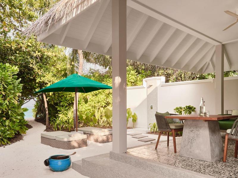 Überdachter Sitzbereich neben einem Sandweg und grünem Sonnenschirm im Garten.