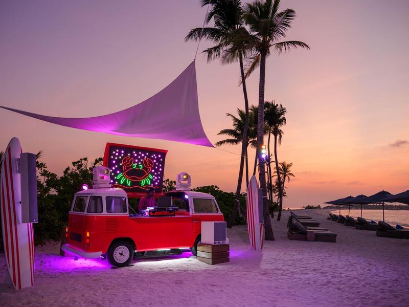 Roter VW-Bus am Strand mit DJ, beleuchtet bei Sonnenuntergang.