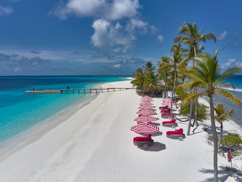 Strand mit weißen Sand, türkisfarbenem Wasser, Palmen und roten Sonnenschirmen.