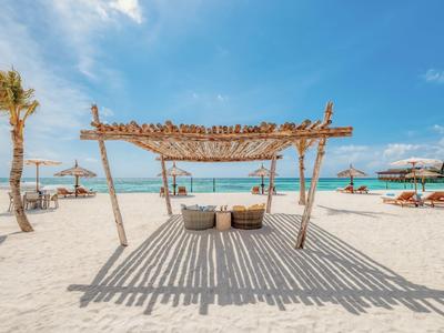 Ein heller Sandstrand mit Palmen, Sonnenschirmen, Sitzgelegenheiten und klarem blauem Wasser unter blauem Himmel.