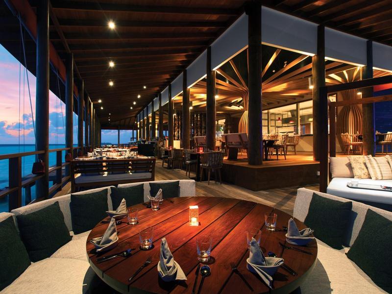 Zewnętrzny obszar jadalny z drewnianymi meblami i widokiem na ocean o zachodzie słońca.