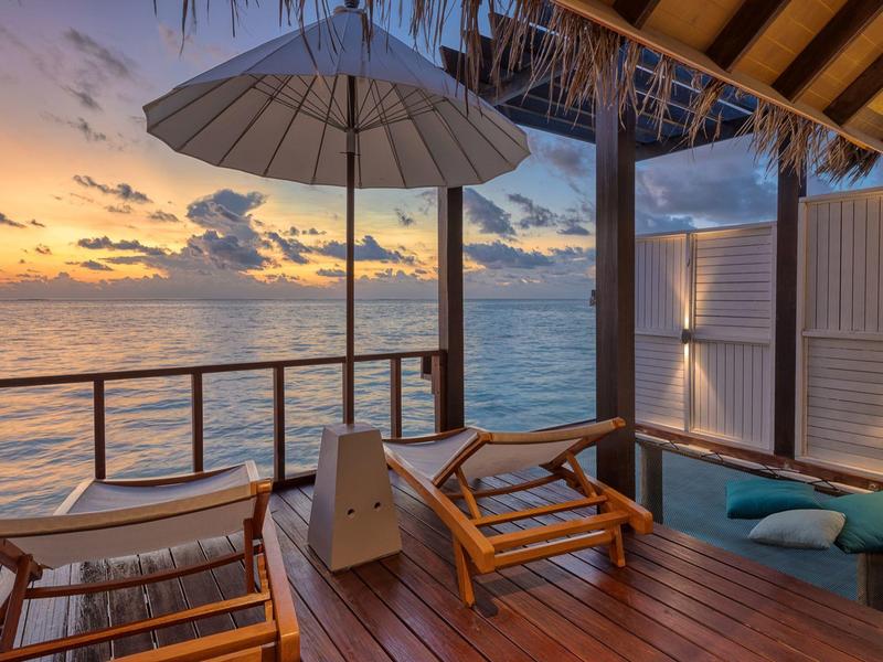 Drewniany taras z leżakami i parasolem z widokiem na ocean o zachodzie słońca.