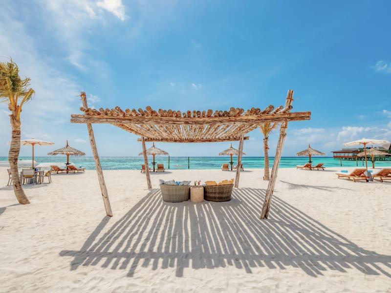 Zacieniona przestrzeń na plaży z wiklinowymi fotelami i parasolami na białym piasku pod błękitnym niebem.