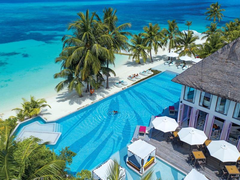 Basen przy plaży z palmami i budynkiem o dachu z trzciny z widokiem na turkusowe morze.