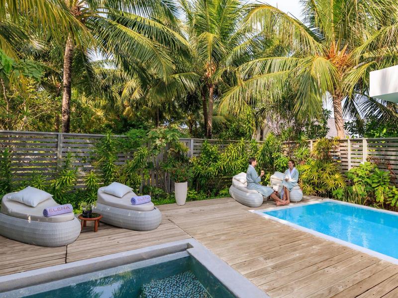 Zewnętrzny obszar basenu z leżakami, drewnianym tarasem i palmami tworzącymi tropikalny klimat.