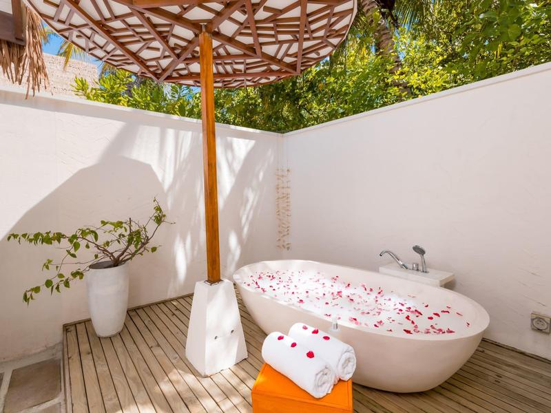 Wanna na zewnątrz z płatkami kwiatów, parasolem i ręcznikami na drewnianym tarasie otoczonym białymi ścianami.