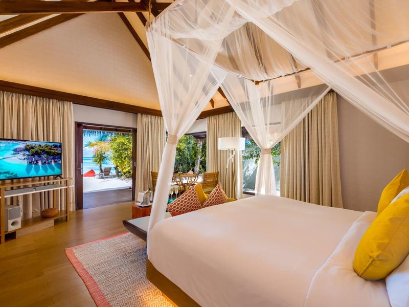 Jasny pokój hotelowy z łóżkiem z baldachimem, żółtymi poduszkami, drewnianą podłogą i telewizorem z widokiem na basen.