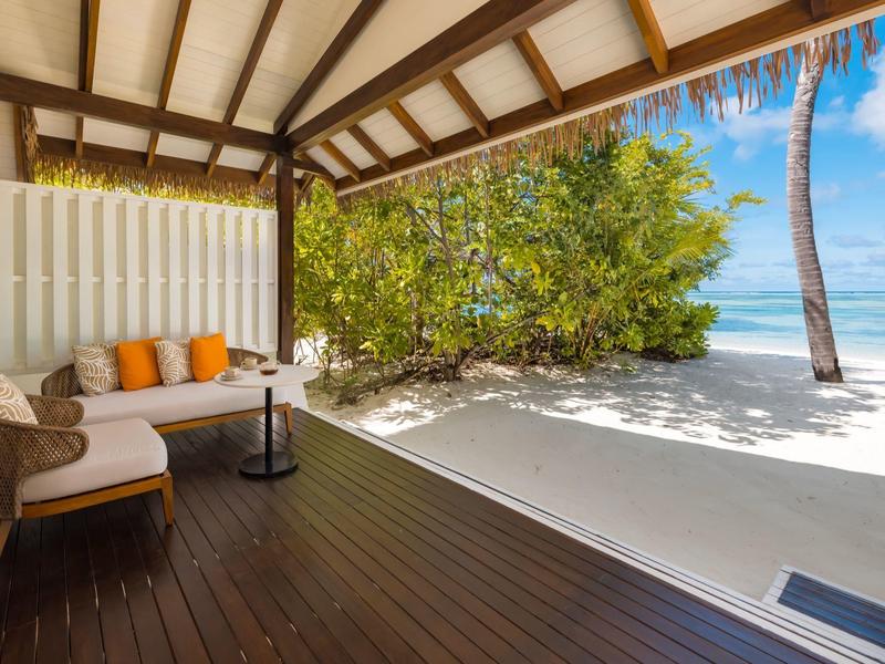 Przytulna kabina przy plaży z drewnianym tarasem, miękkimi siedziskami, palmami i białym piaskiem.