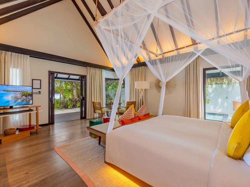 Jasny pokój hotelowy z łóżkiem z baldachimem, drewnianą podłogą, telewizorem i otwartymi drzwiami na ogród.