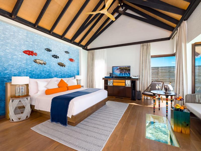 Jasny pokój hotelowy z drewnianą podłogą, dużym łóżkiem, kolorową sztuką na ścianie i widokiem na morze.