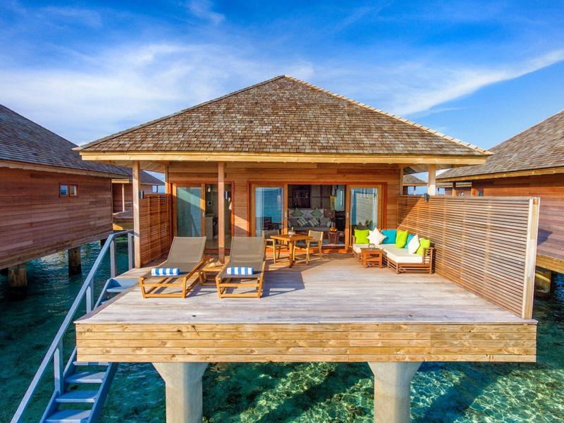 Bungalow sull'acqua con terrazza in legno, lettini e zona salotto accogliente sotto un cielo azzurro.