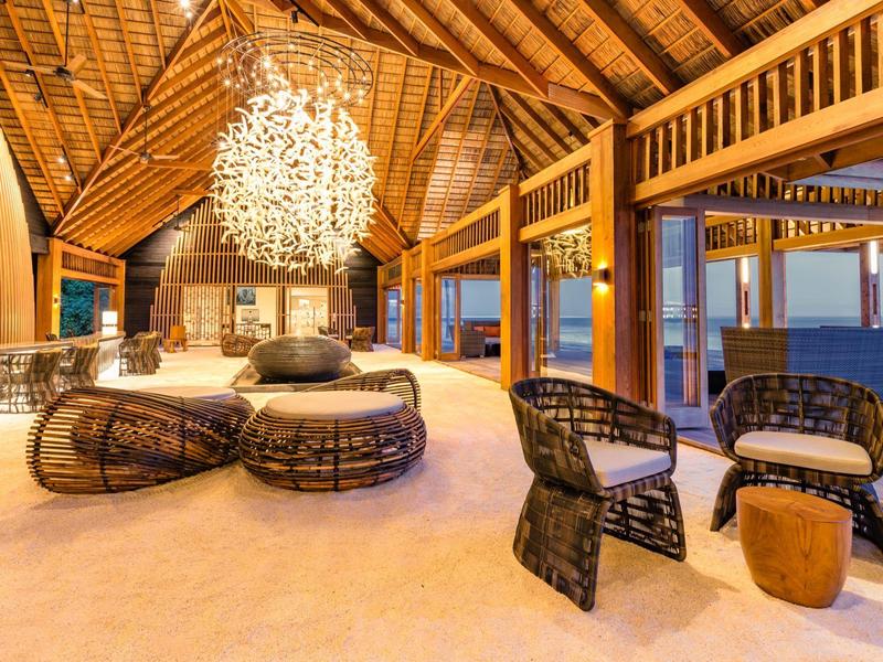 Lounge aperta di lusso con mobili in rattan e imponente lampadario sulla spiaggia sabbiosa.