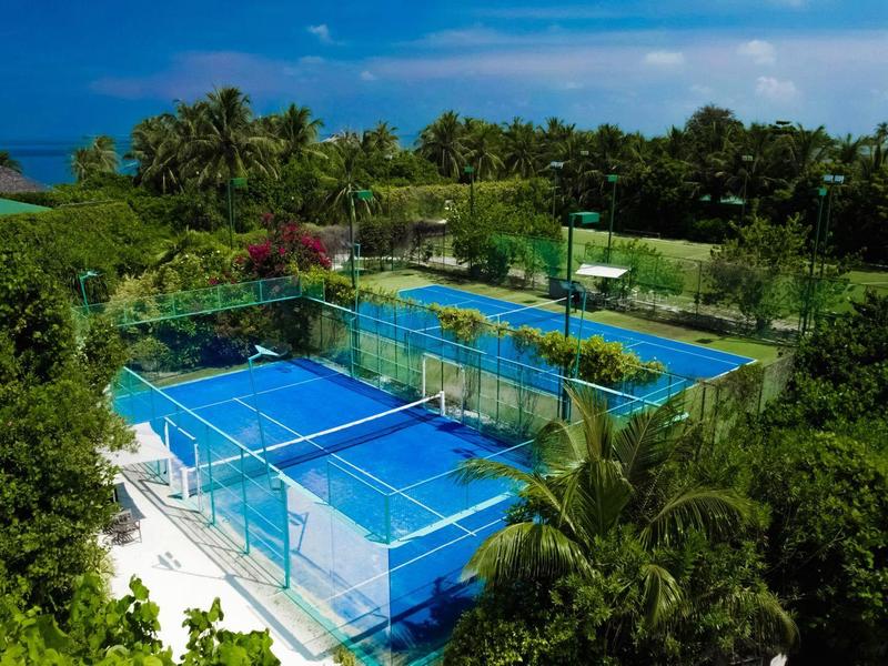 Diverse corti da tennis blu circondati da una vegetazione lussureggiante e palme in un ambiente tropicale.