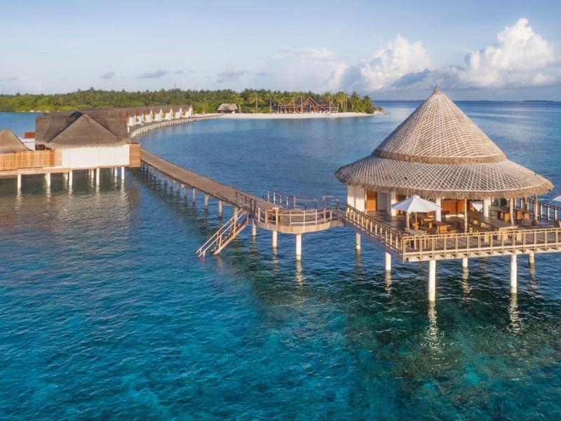 Bungalows sobre pilotes con restaurante sobre agua turquesa junto a una isla tropical.