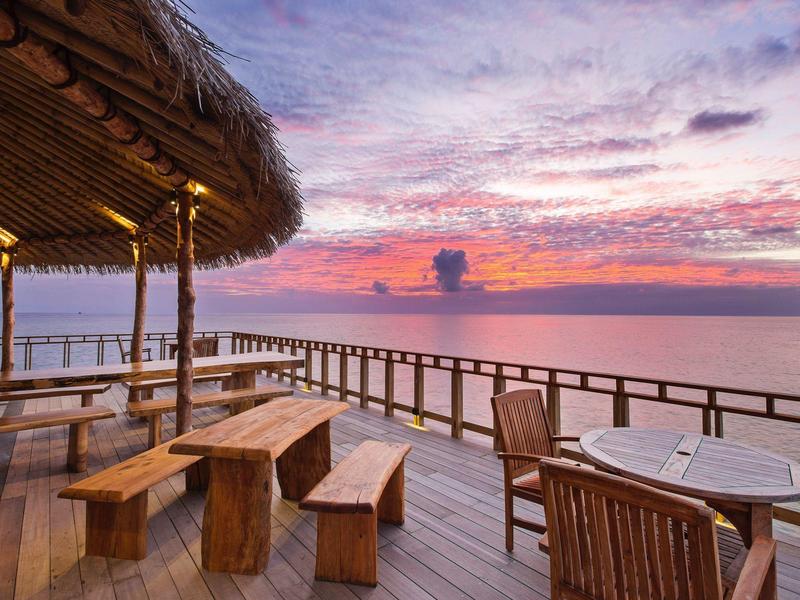 Terraza de madera con áreas de asiento y vista del atardecer sobre el mar tranquilo.