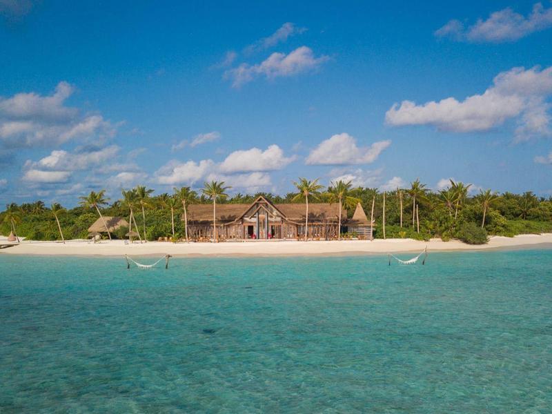 Pequeña isla tropical con playa de arena, palmeras y agua clara turquesa bajo un cielo azul.
