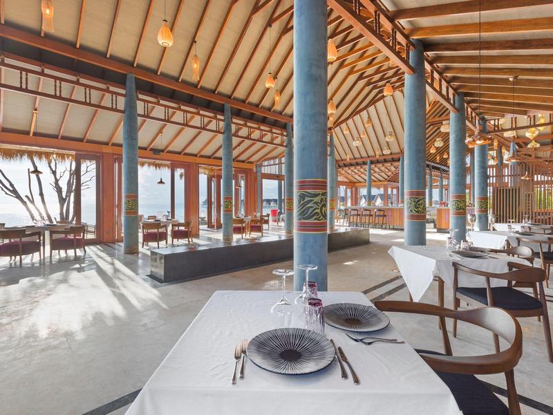 Restaurante de hotel luminoso y abierto con techo de madera, mesas blancas y vista al mar a través de grandes ventanas.
