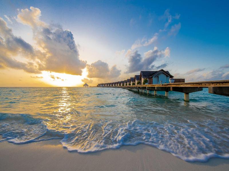 Atardecer en la playa con bungalows sobre un muelle y olas suaves en la orilla de arena.