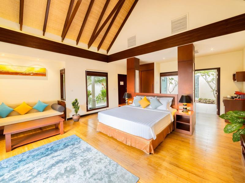 Habitación de hotel luminosa con suelo de madera, cama doble, sofá, alfombra y grandes ventanas con vista al verde.