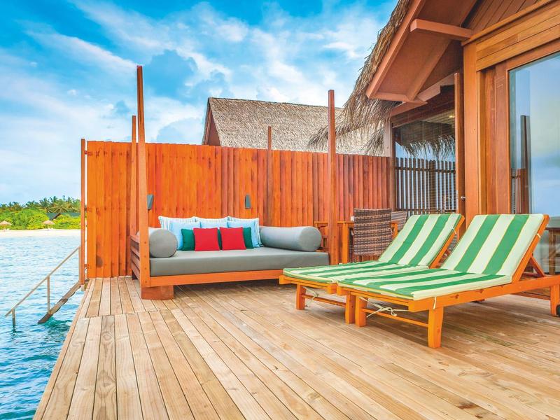 Terraza de madera con tumbonas y sofá junto al agua, conectada a un bungalow de hotel.