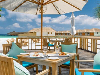 Terrasse mit Holztischen und blauen Kissen, Blick auf Meer und Strandhäuser bei sonnigem Wetter