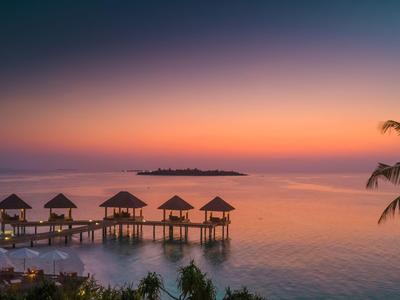 Sonnenuntergang über Bungalows auf Stelzen im Meer mit Palmen im Vordergrund.
