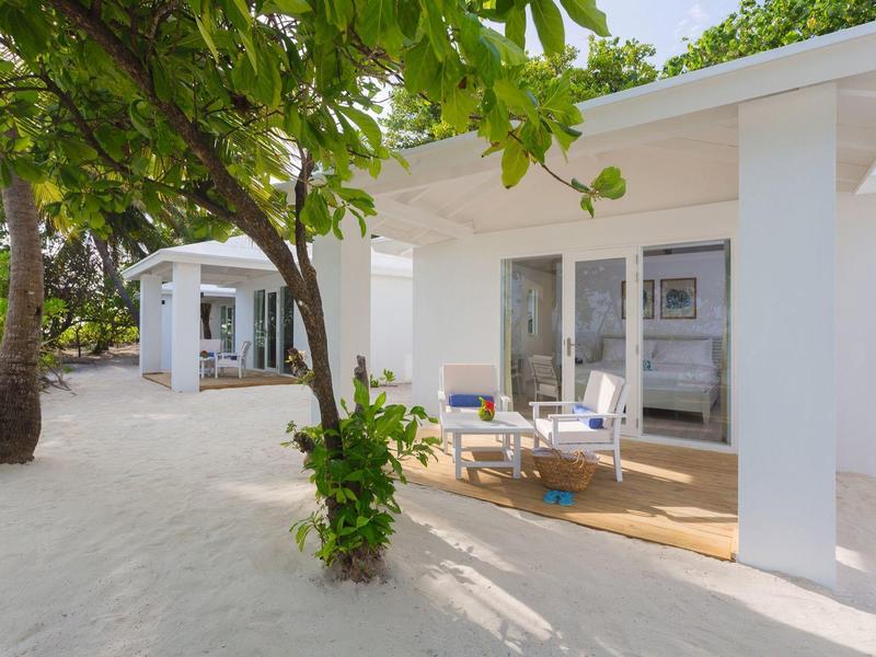 Weiße Bungalows mit Veranden und Gartenmöbeln, umgeben von Sand und grünen Bäumen.