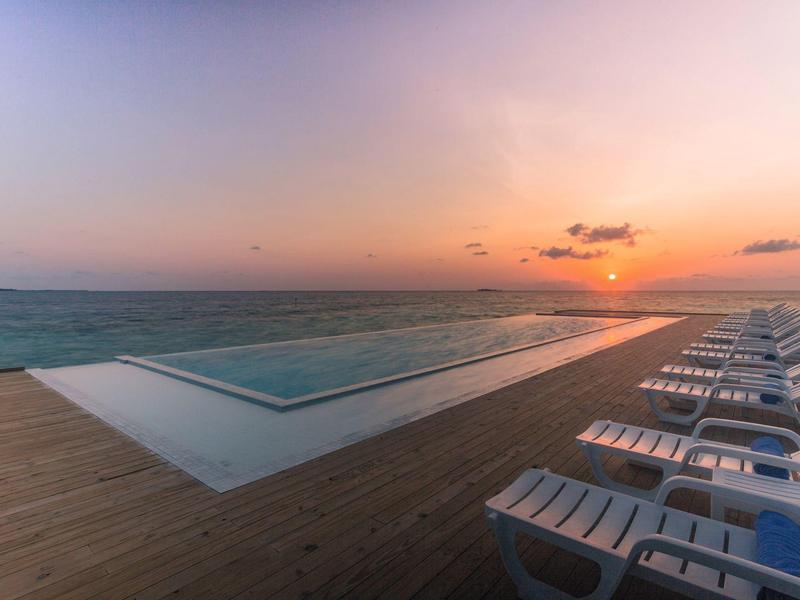 Infinity-Pool mit weißen Liegestühlen auf Holzdeck bei Sonnenuntergang am Meer.