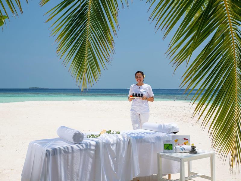Massage-Tisch mit Handtüchern am Strand unter Palmen, Frau im Hintergrund mit Spa-Ausstattung.