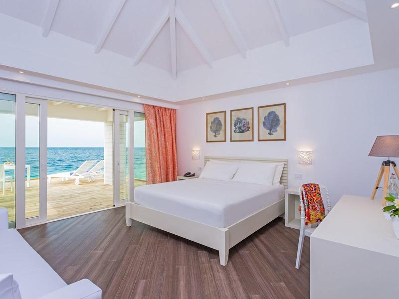 Helles Hotelzimmer mit Doppelbett, Holzfußboden und Meerblick durch große Glasfenster.