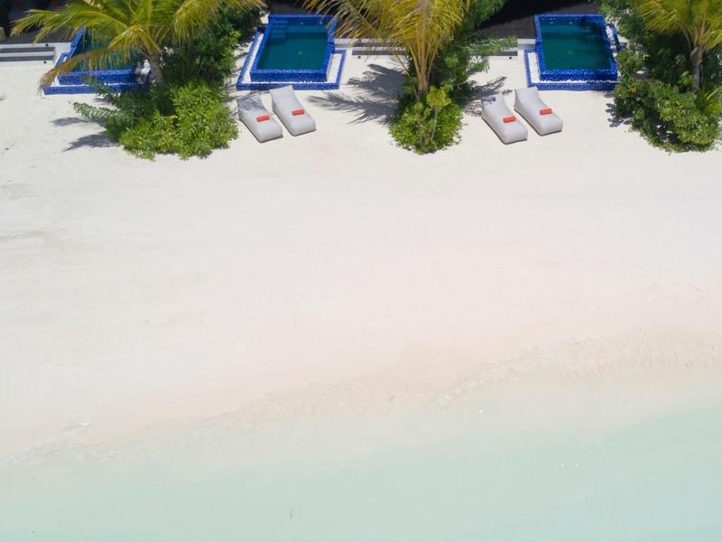 Trois chaises longues sous des palmiers près de toits de chaume sur une plage de sable blanc avec eau turquoise.