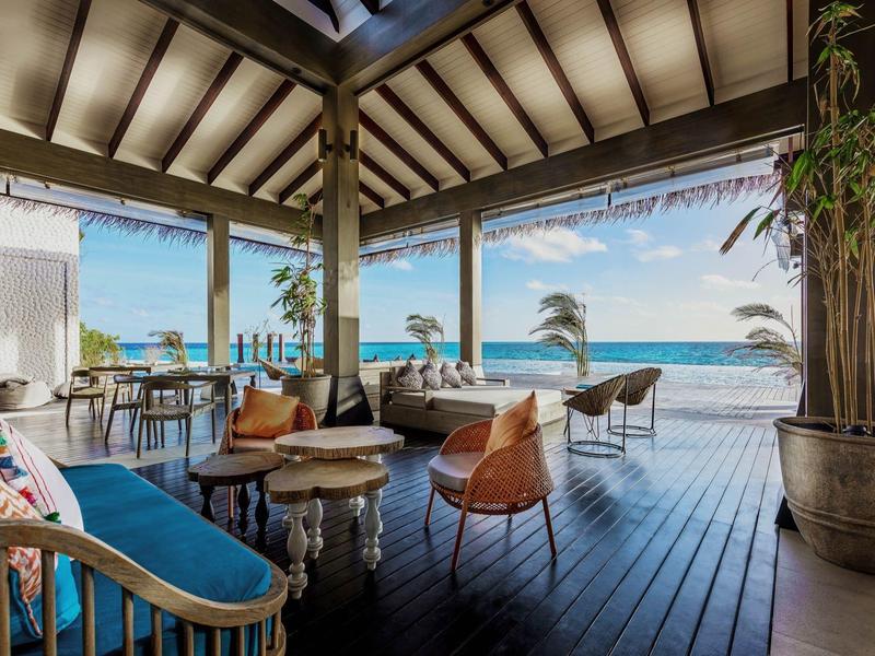 Espace lounge ouvert avec terrasse en bois, canapé, chaises et vue sur la mer.