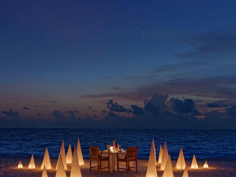 Dîner sur la plage avec une table dressée entourée de cônes lumineux au coucher du soleil.
