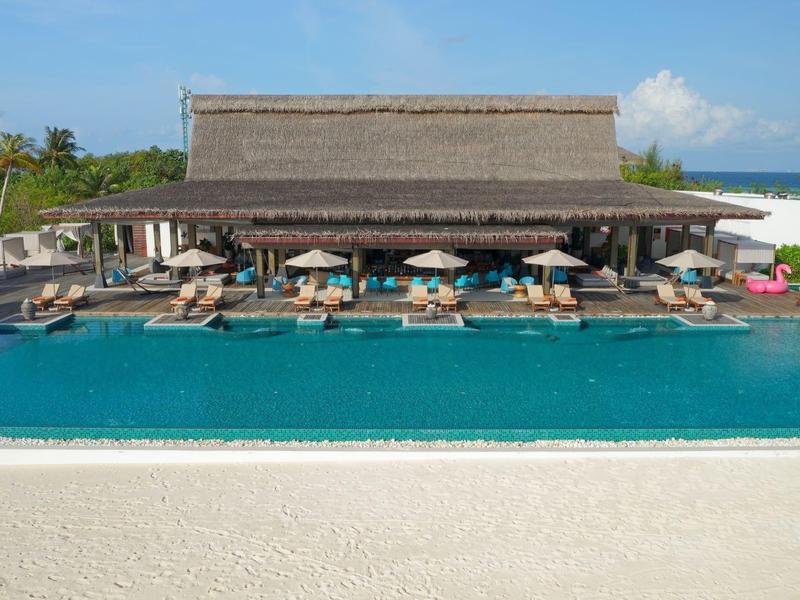 Grande piscine d'hôtel avec chaises longues et parasols devant un bâtiment à toit de chaume ouvert dans la station balnéaire.