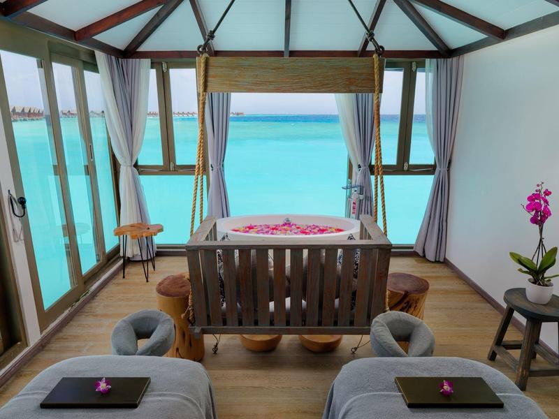 Chambre d'hôtel avec vue sur l'eau turquoise, deux lits et une table en bois avec des fleurs.