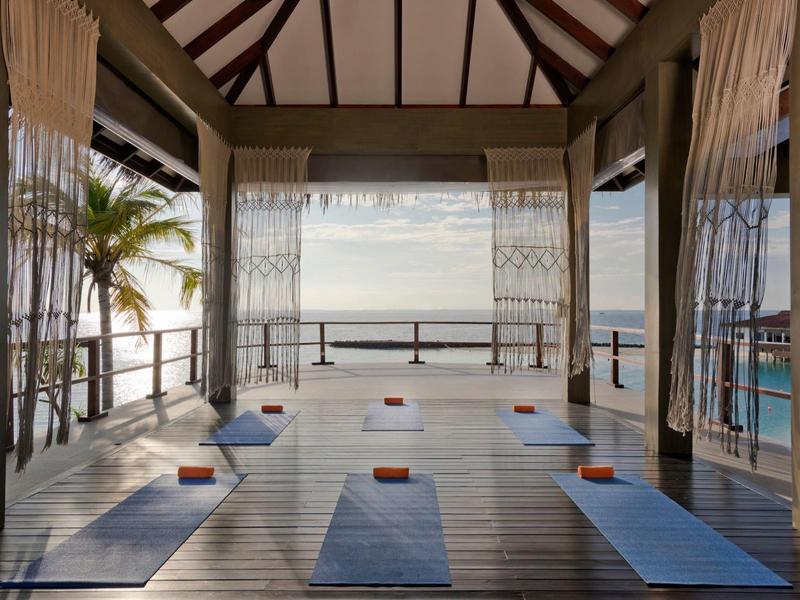 Studio de yoga avec tapis, ouvert sur la mer et terrasse en bois avec rideaux.