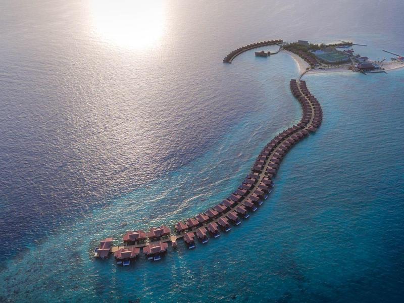 Vue aérienne de bungalows sur pilotis s'étendant le long d'une jetée courbée au-dessus d'une mer turquoise.