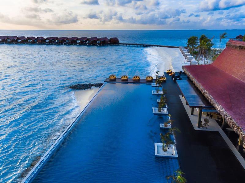Piscine à débordement avec vue sur la mer et bungalows sur l'eau sous un ciel nuageux.