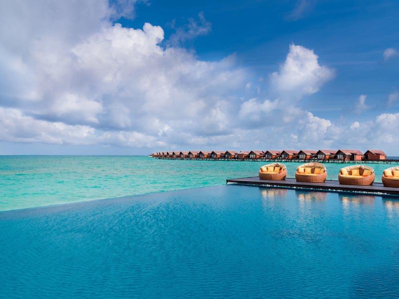 Piscine à débordement avec vue sur la mer turquoise et des bungalows sous un ciel bleu.